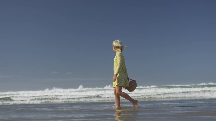 A blonde female child in a yellow dress walking through the tide on the beach carrying a wicker basket