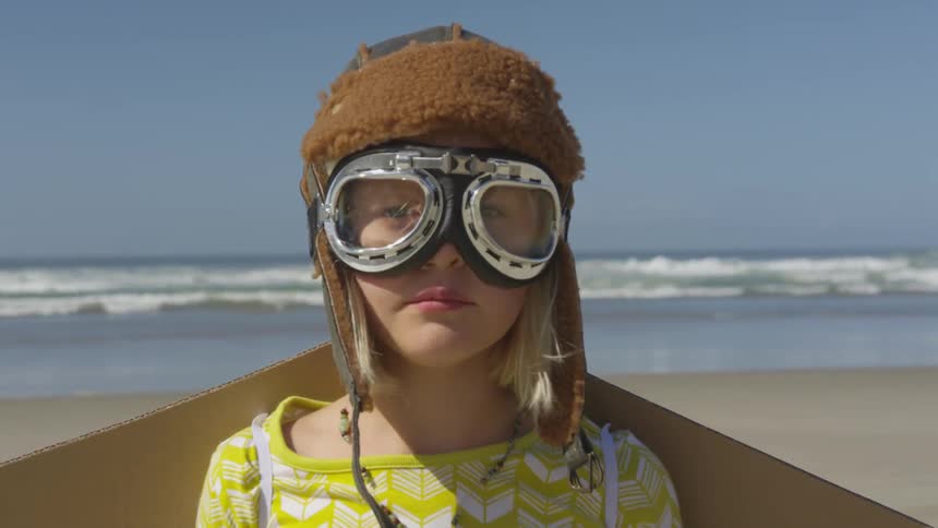 A girl on the beach with a hat and goggles on and cardboard wings strapped to her back