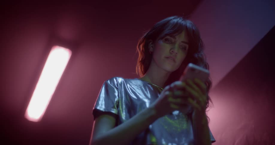 A woman in a shiny silver shirt uses a smartphone beneath a fluorescent light