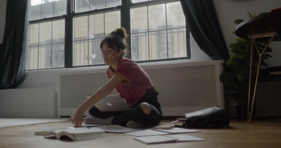 A woman sits on the floor typing on a laptop and looking at books