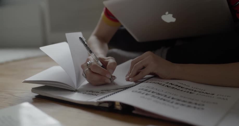 A person sits cross legged on the floor while writing notes in a notebook while a mac sits on their lap