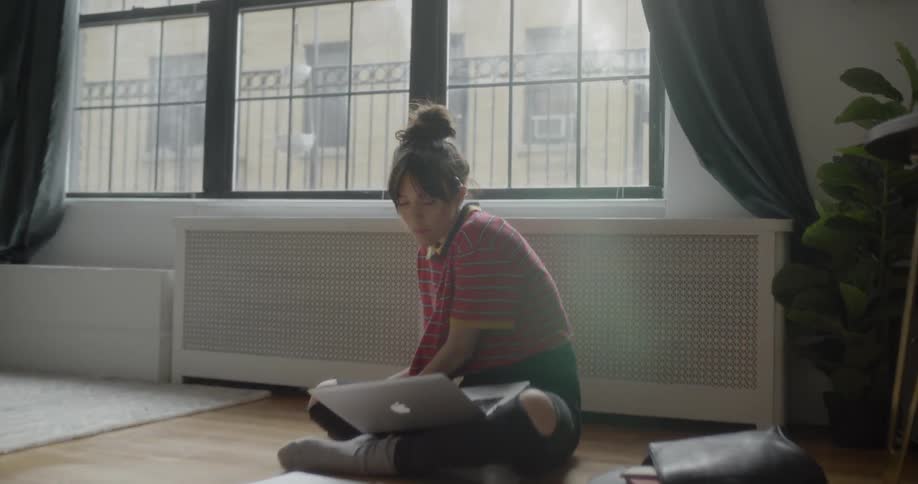A woman sits on the floor of a bedroom eating and working on a laptop