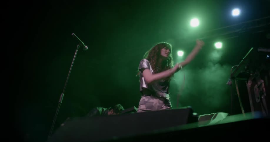 A woman with wavy hair and a glossy crop top sings into a microphone and dances on stage accompanied by other musicians