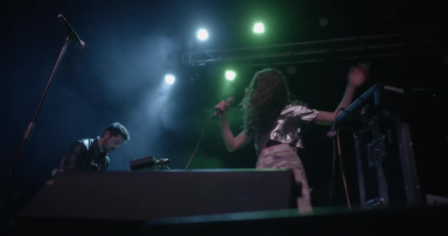 A woman on stage holding a mic and singing while a guy in the background is on keyboards