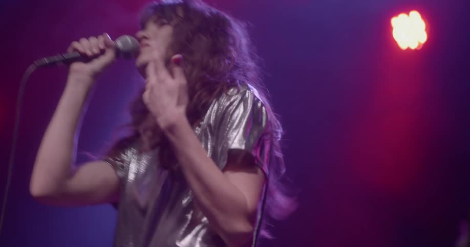 A woman with wavy hair and a glossy crop top sings into a microphone and dances on stage