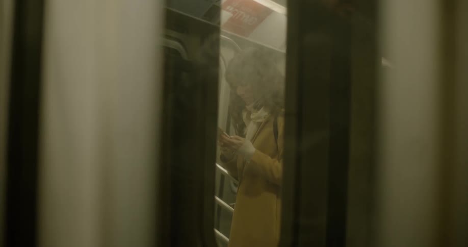 A woman in a mustard color coat standing in a carriage on the metro answers her phone