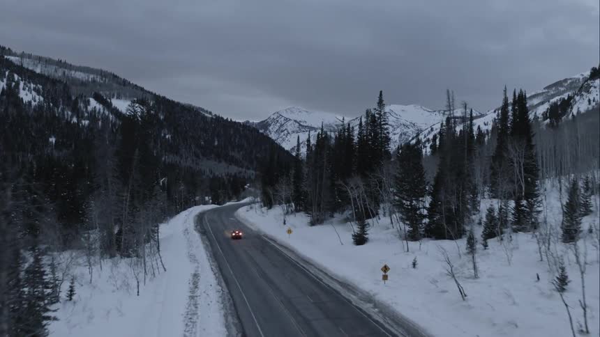 A car drives on an icy mountain road flanked by snowy mountains and trees