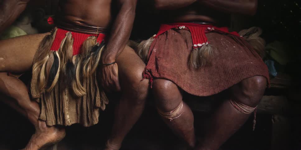 Two men wearing loincloths sit on a bench as one of the men adjusts his loincloth