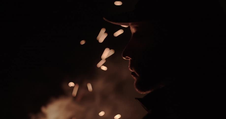 Man who wears cap sits down in front of sparkling fire at night