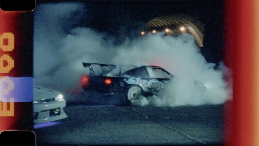 A stock car with an aerofoil spins its wheels to create smoke in front of an aircraft hangar at night