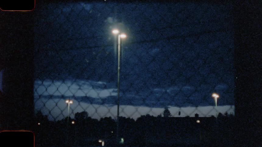 Street lamps and cars parked in an almost empty lot seen through a chain link fence at night