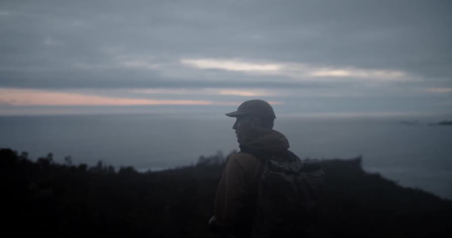 A hiker standing on a hill near a the ocean on a cloudy evening
