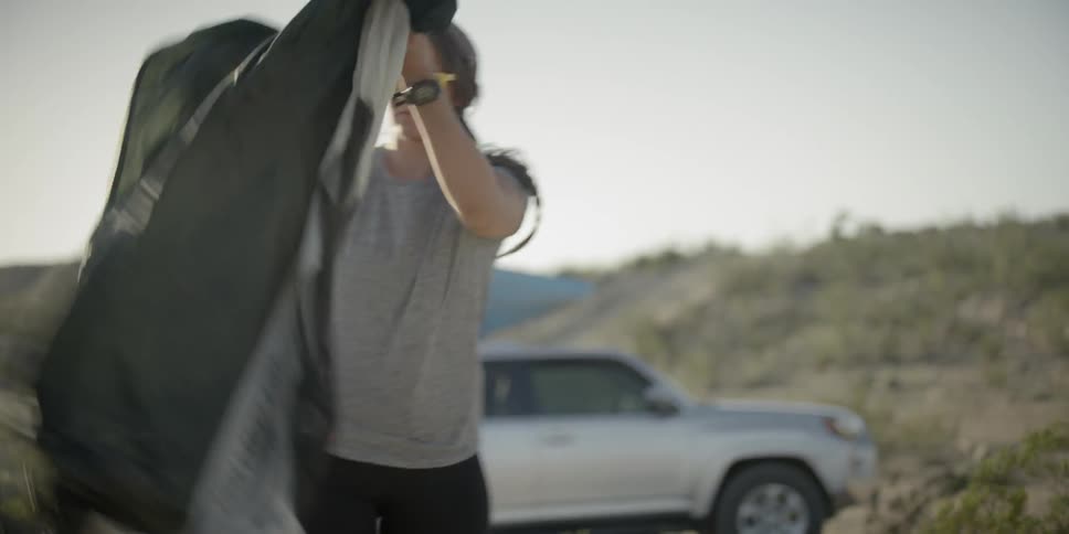A woman opens up her tent in an arid environment while her suv is parked behind her with an open trunk