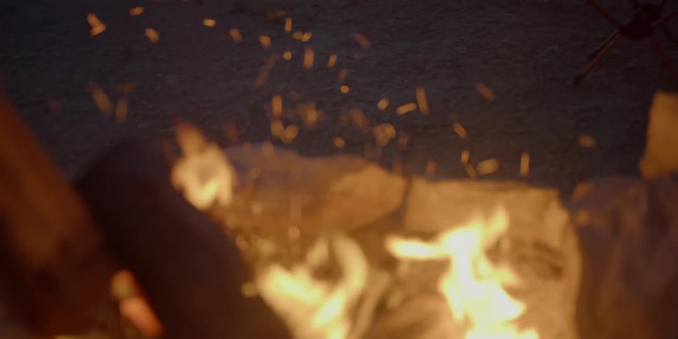 A person agitating a log on a bonfire while another person in the background sits in a camping chair