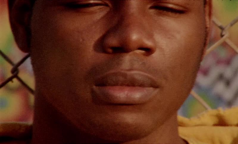 A young black man leans against a chain link fence