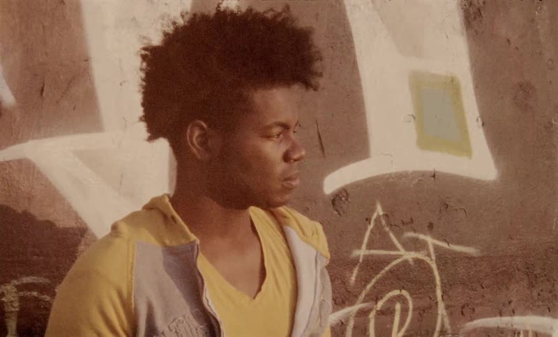 A black teenager stands in front of a wall with graffiti on it