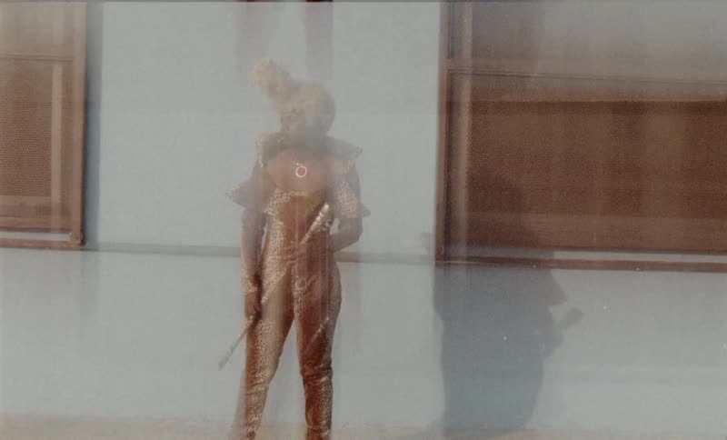 A black man dressed in an outlandish costume walks around outside a building