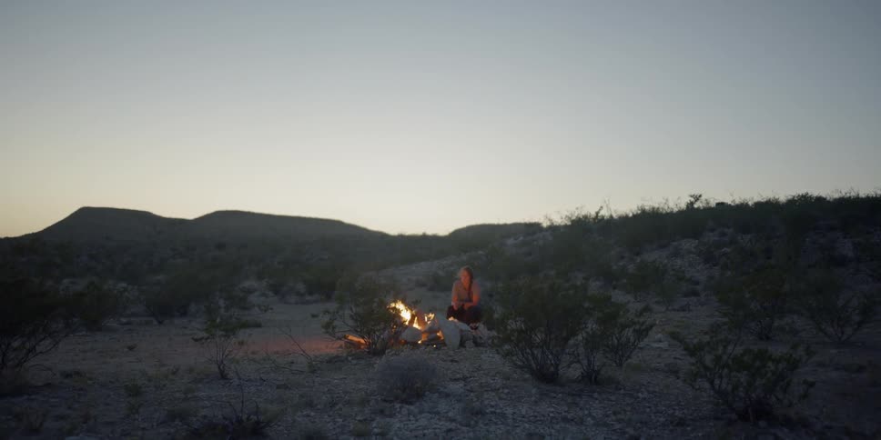 Woman crouching alone in front of fire on the land warms up at dusk