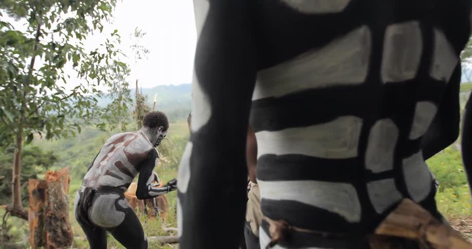 A group of men painted like skeletons, gather around as some of the men apply paint to the other me