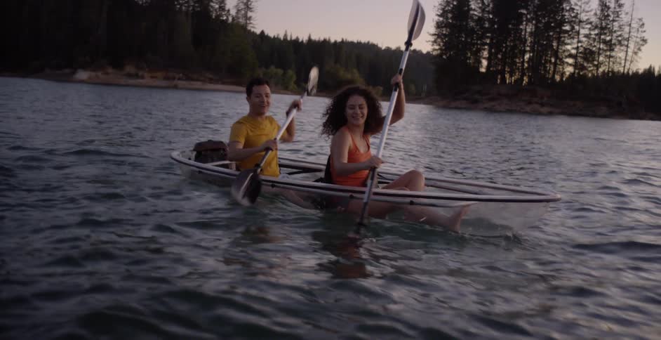 A woman and a man rowing a transparent canoe through a lake in the evening