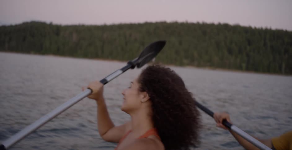 A woman and a man using oars to paddle a boat through a lake in the evening