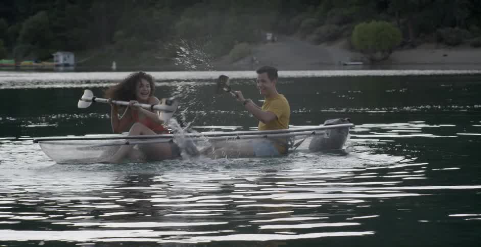 A man and woman in a clear kayak splashing water and laughing