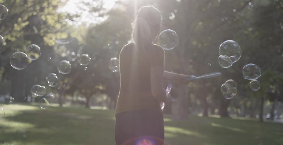 A blonde woman pays with bubbles and spins in a circle while smiling under sunlight
