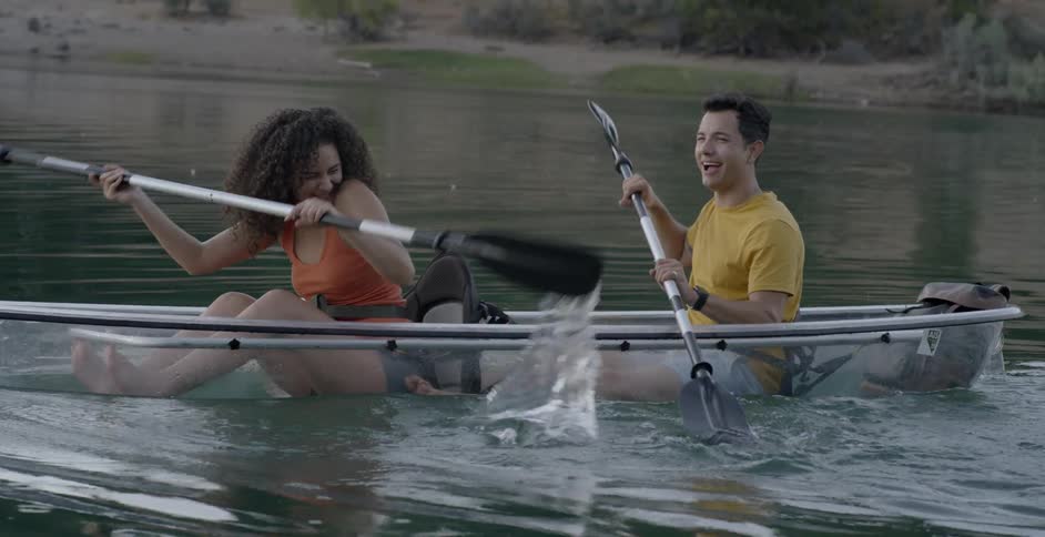A man and a woman sit in a row boat and splash each other with the oars
