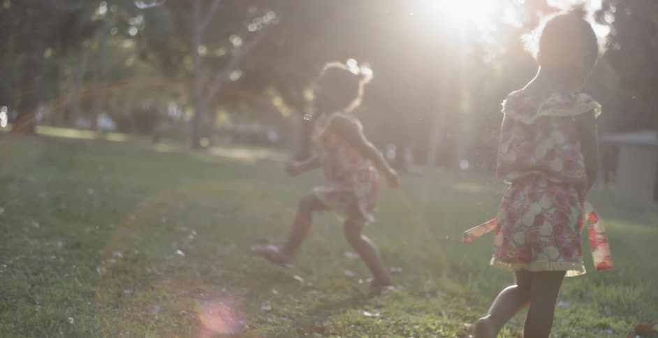 Two young children in identical clothes play with bubbles on a lawn at low sun