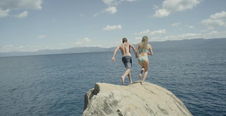 A couple holds hands as they run and jump off a rock into the ocean below