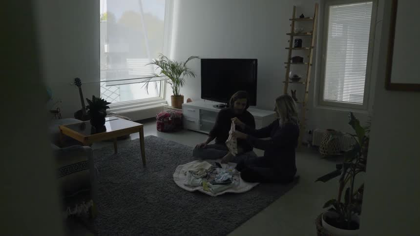 A couple folds baby clothes while sitting on a gray rug on the floor in a living room