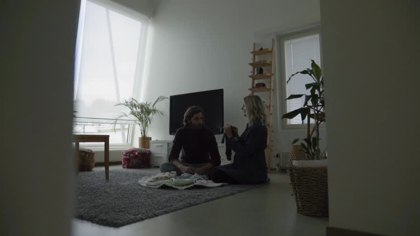 A pregnant woman kneels on the living room rug next to her husband holding up baby clothes