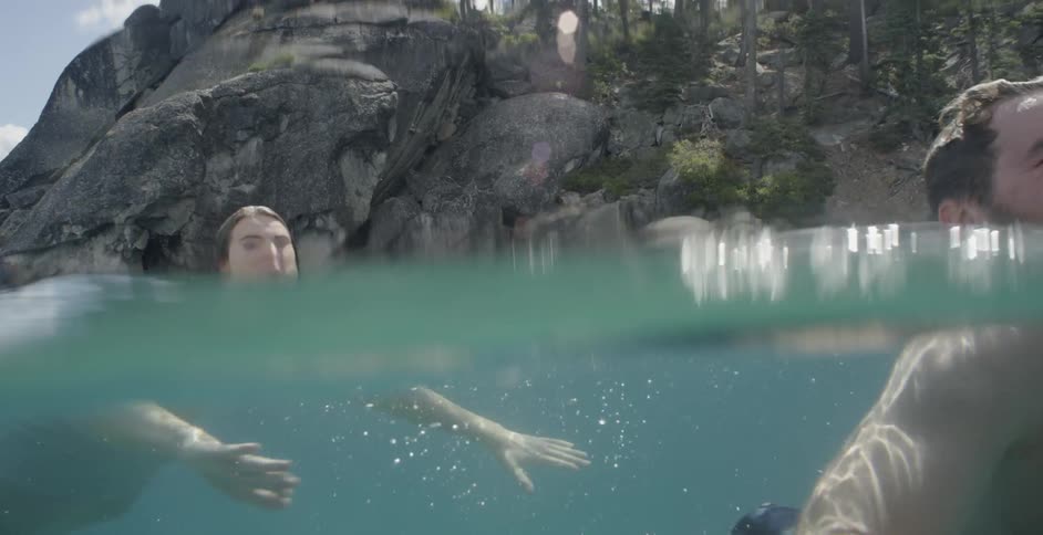A man and woman swim in clear water with cliffs trees and rocks in the distance