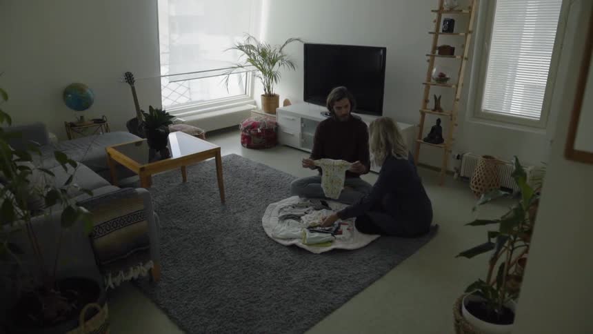 A young couple look at baby clothes while sitting on a rug in their living room