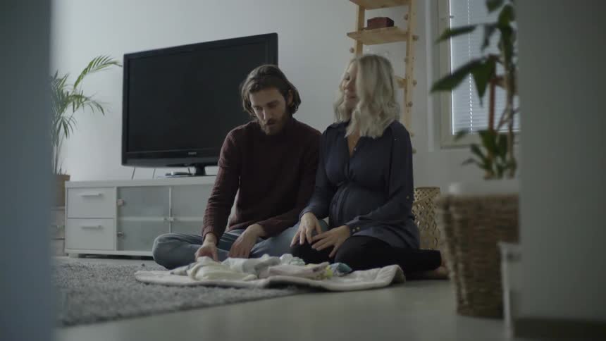 A man and a woman sitting on a carpet while looking at baby clothing on a cushion