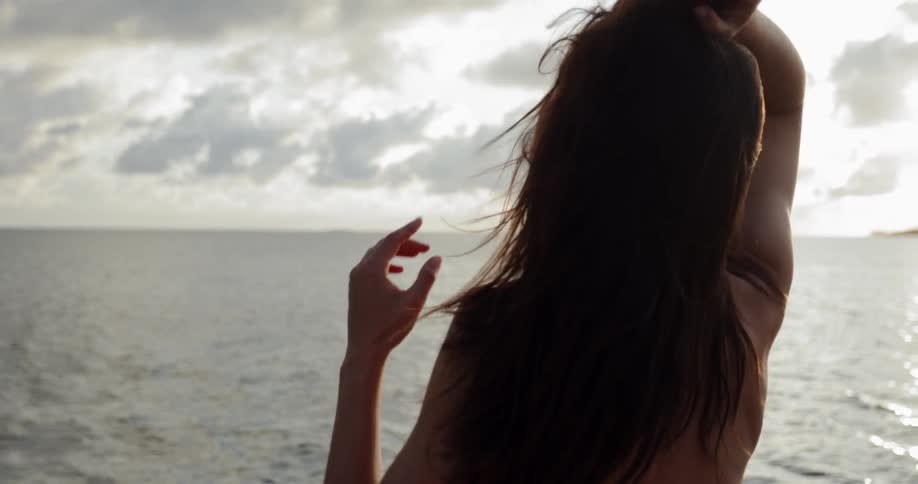 A woman wearing a bathing suit flips and fluffs her long brown hair near the ocean