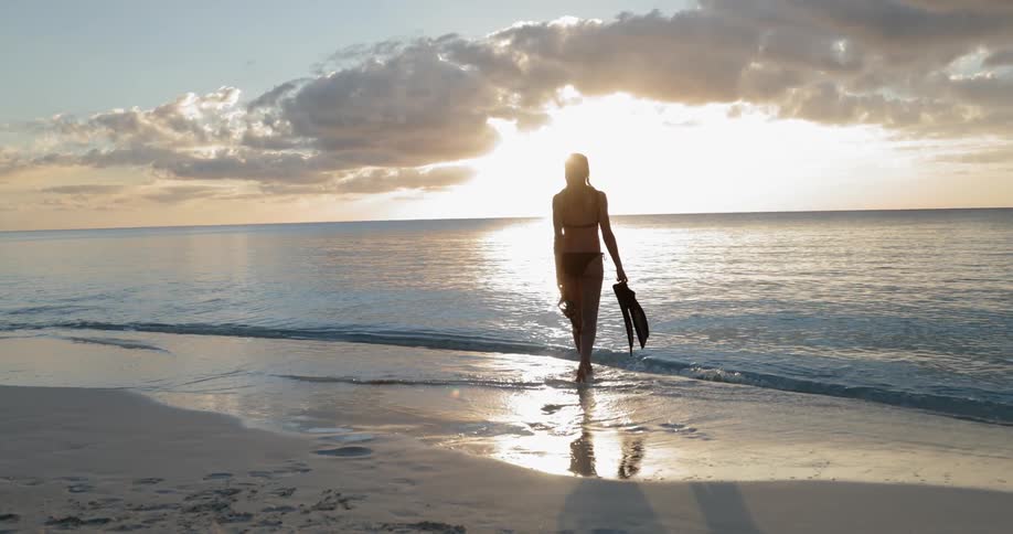 A bikini clad woman walking towards the sea with flippers in one hand and snorkel in the other at sunset