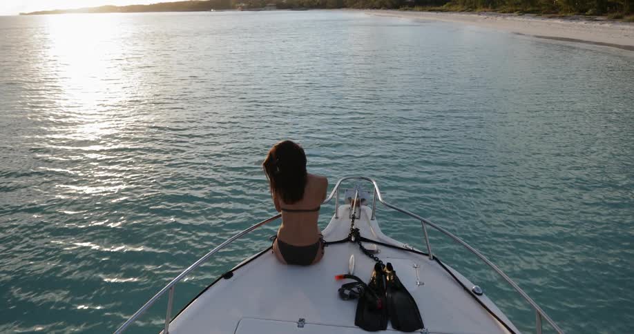 A woman in a bikini sitting on the deck of a boat dangling her legs off the front