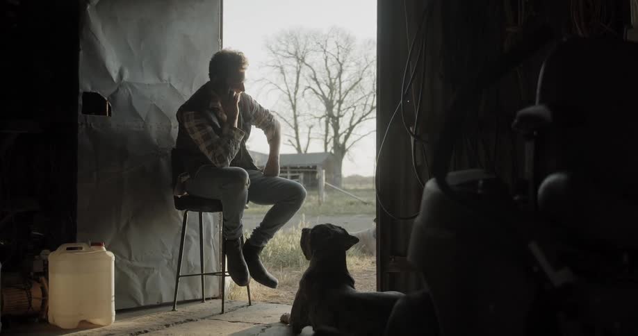 A dog looks up at his master who is sitting on a stool talking on the phone in the doorway of a work shed