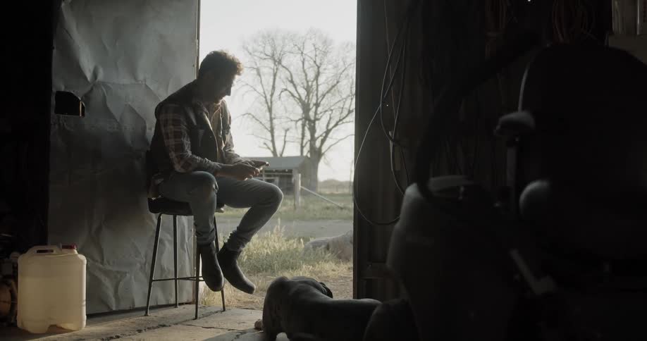 A man is texting on his phone while sitting on a stool in an open doorway of a barn