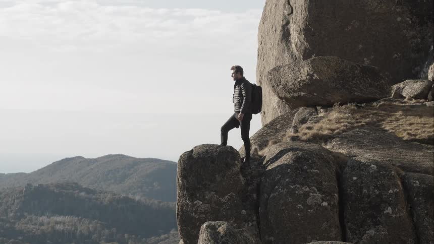 A man wearing a backpack stands with one leg raised on the edge of a rocky ridge