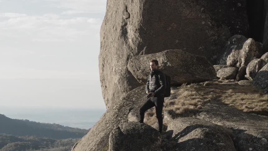 Man wearing black backpack and coat stands on the rock and takes picture of the landscape during daylight