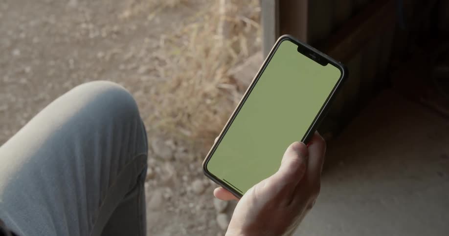 A man sitting in the doorway of a shed holds a smartphone in his hand