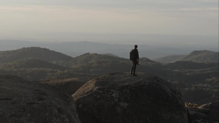 A male hiker standing on a boulder at high altitude and looking down over the countryside