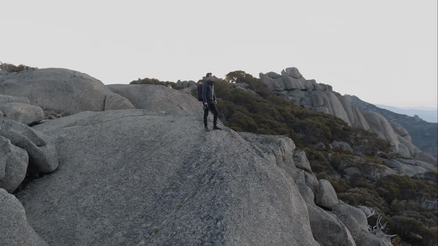 Man wearing backpack and holding camera stands on a big rock and looks at the horizon at sunset
