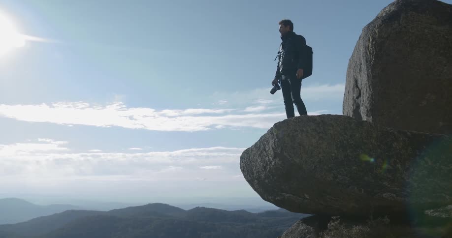 A man climbs onto a large boulder and takes a photo of the scenery