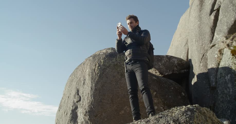 A white male wearing a backpack stands atop a large rock to take a picture