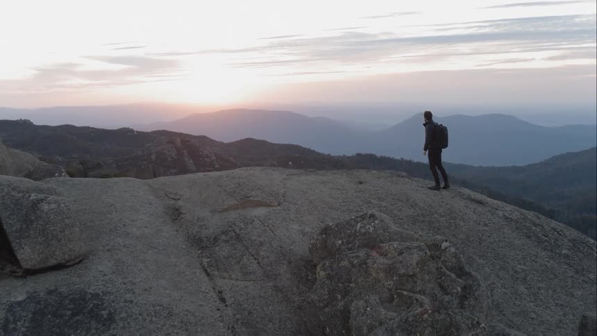 A white man stands atop a mountain and takes in a view of a valley