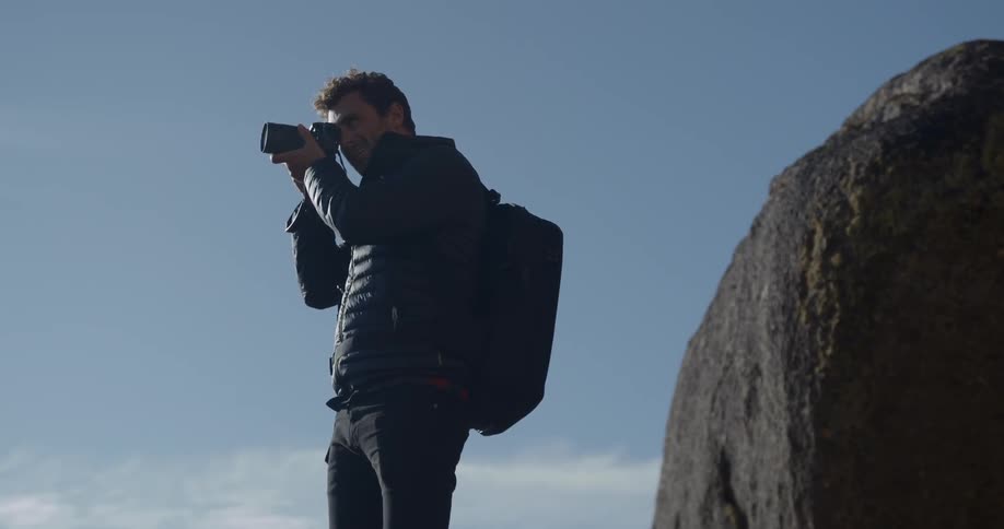 Man wearing black backpack and coat takes picture next to a big rock during daylight
