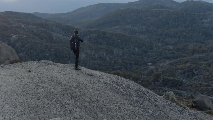 Aerial shot of a man standing on a cliff looking at the terrain with a camera at sunset or sunrise
